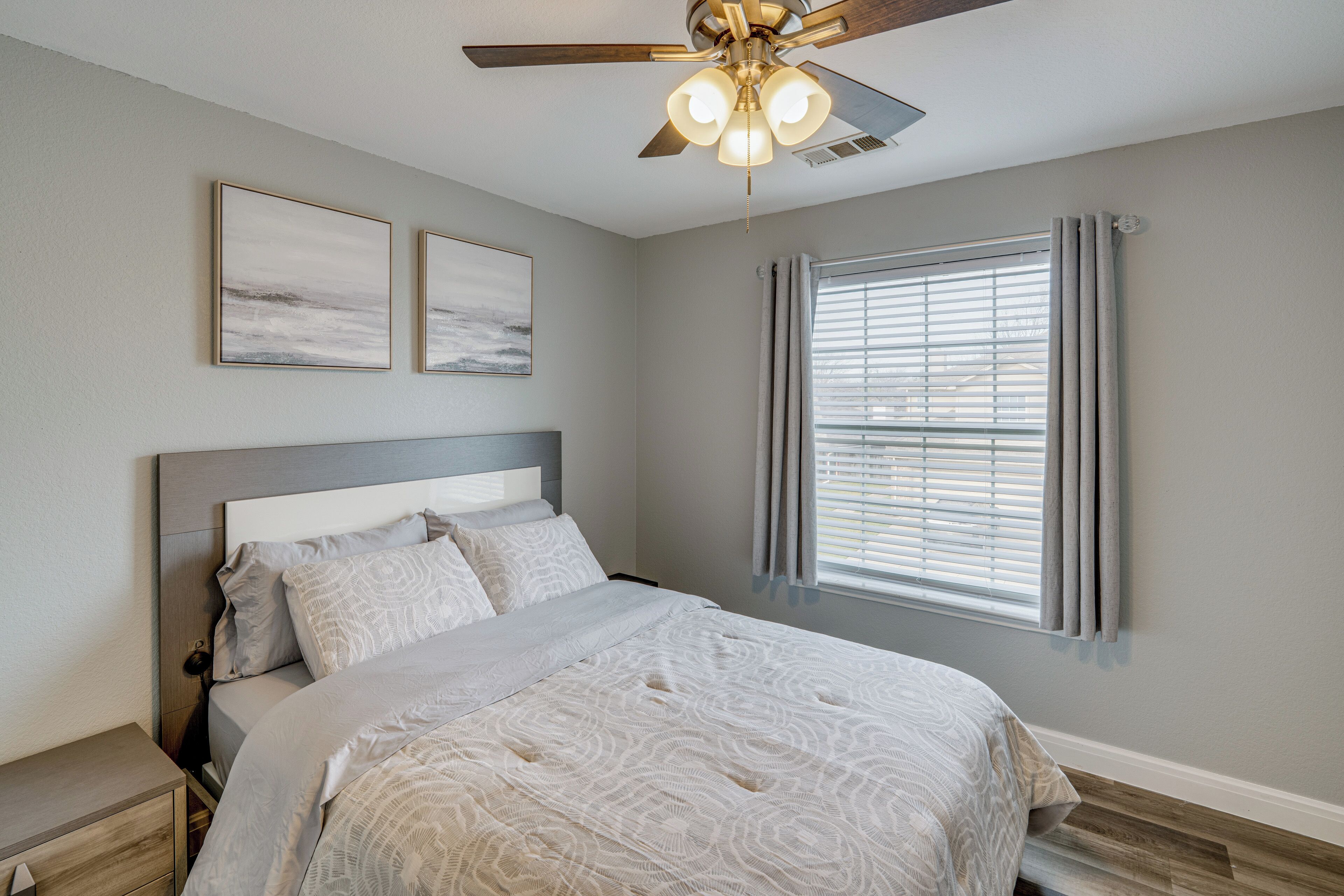 Une chambre à coucher comprenant un lit, un ventilateur de plafond, une fenêtre avec des stores et deux tableaux accrochés au mur.