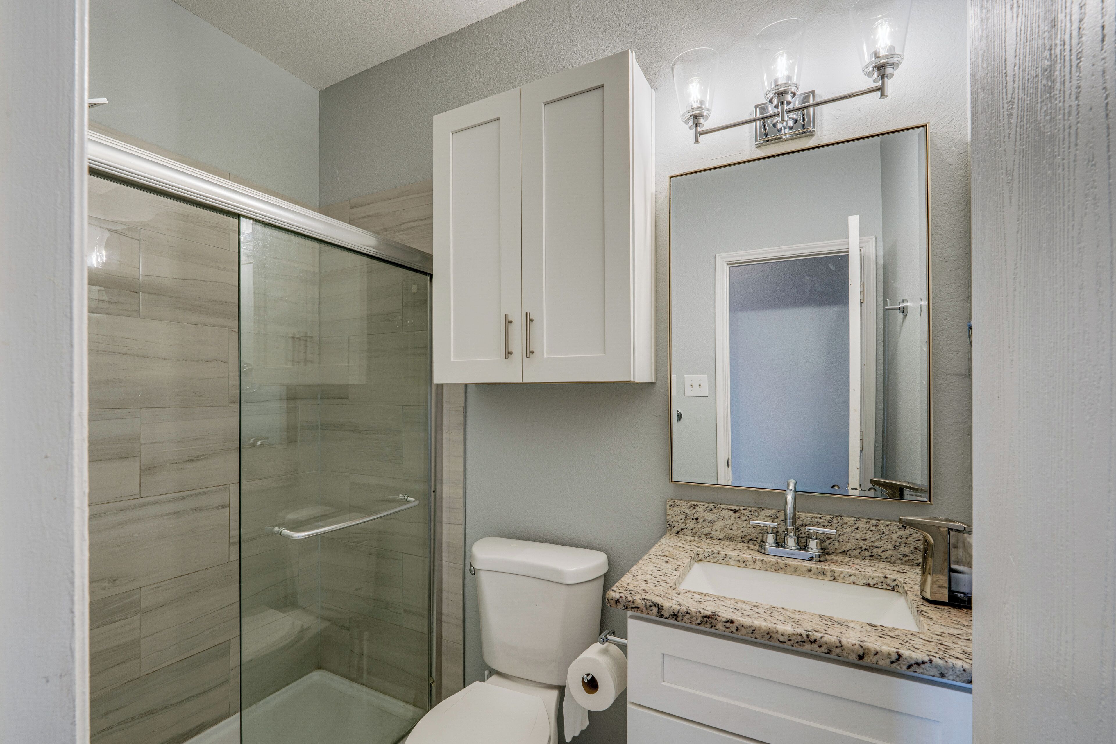 A bathroom with a glass shower enclosure, a toilet, a sink with a granite countertop, and a mirror above the sink.