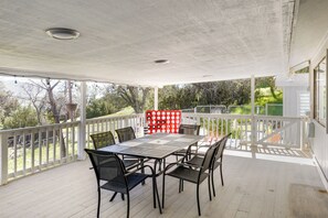 A covered outdoor patio with a table and chairs, a white railing, and a view of trees and a fence.