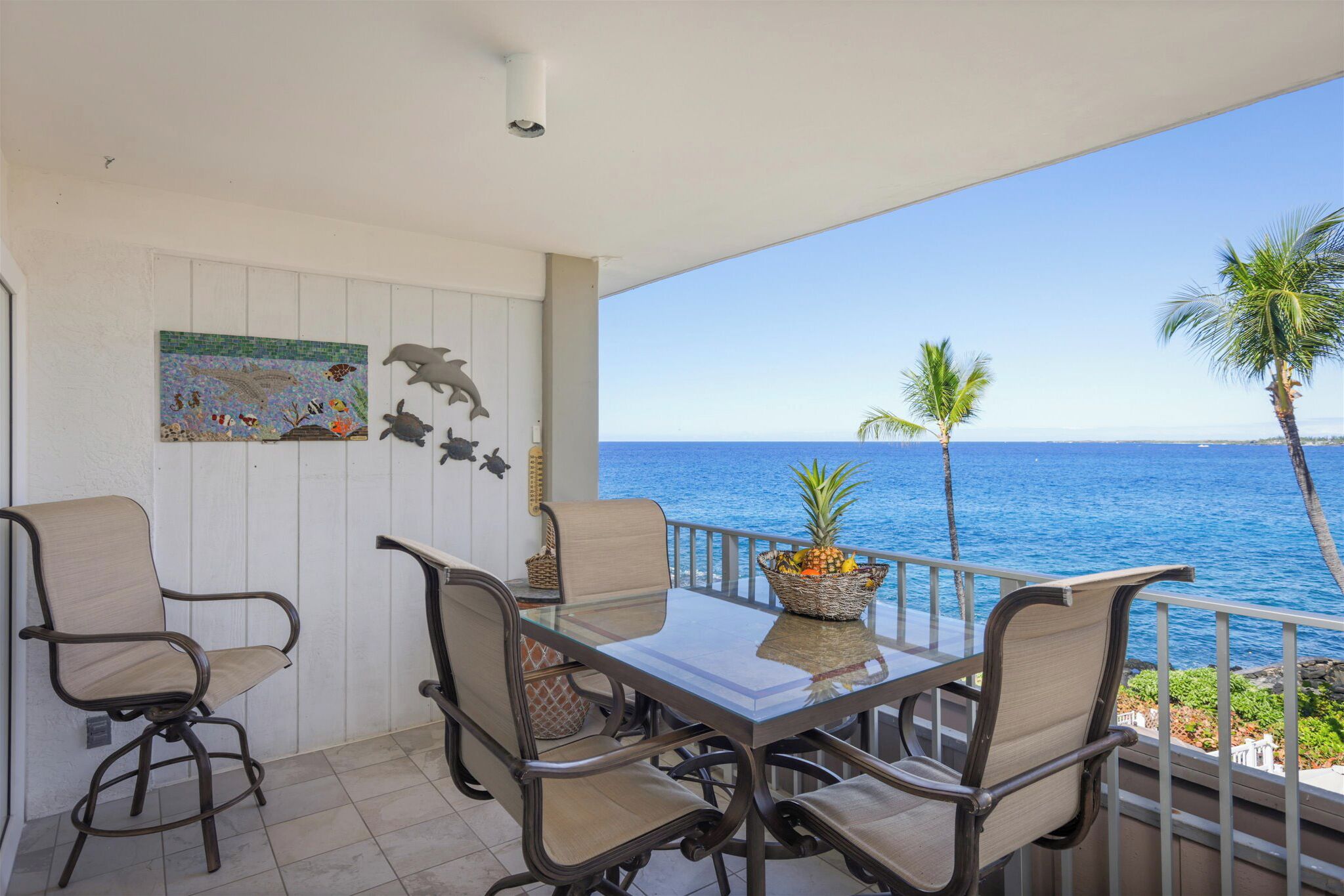 A balcony with a glass table, four chairs, a decorative plant, and a view of the ocean.
