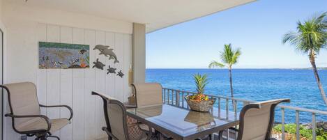 A balcony with a glass table, four chairs, a decorative plant, and a view of the ocean.