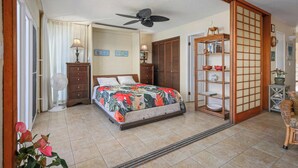 A bedroom with a bed, a ceiling fan, a fan, a dresser, a shelf with decorative items, and a sliding door. - Top Floor Oceanfront Condominium in Kailua-kona (Kailua-Kona)