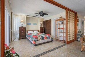 A bedroom with a bed, a ceiling fan, a fan, a dresser, a shelf with decorative items, and a sliding door. - Top Floor Oceanfront Condominium in Kailua-kona (Kailua-Kona)