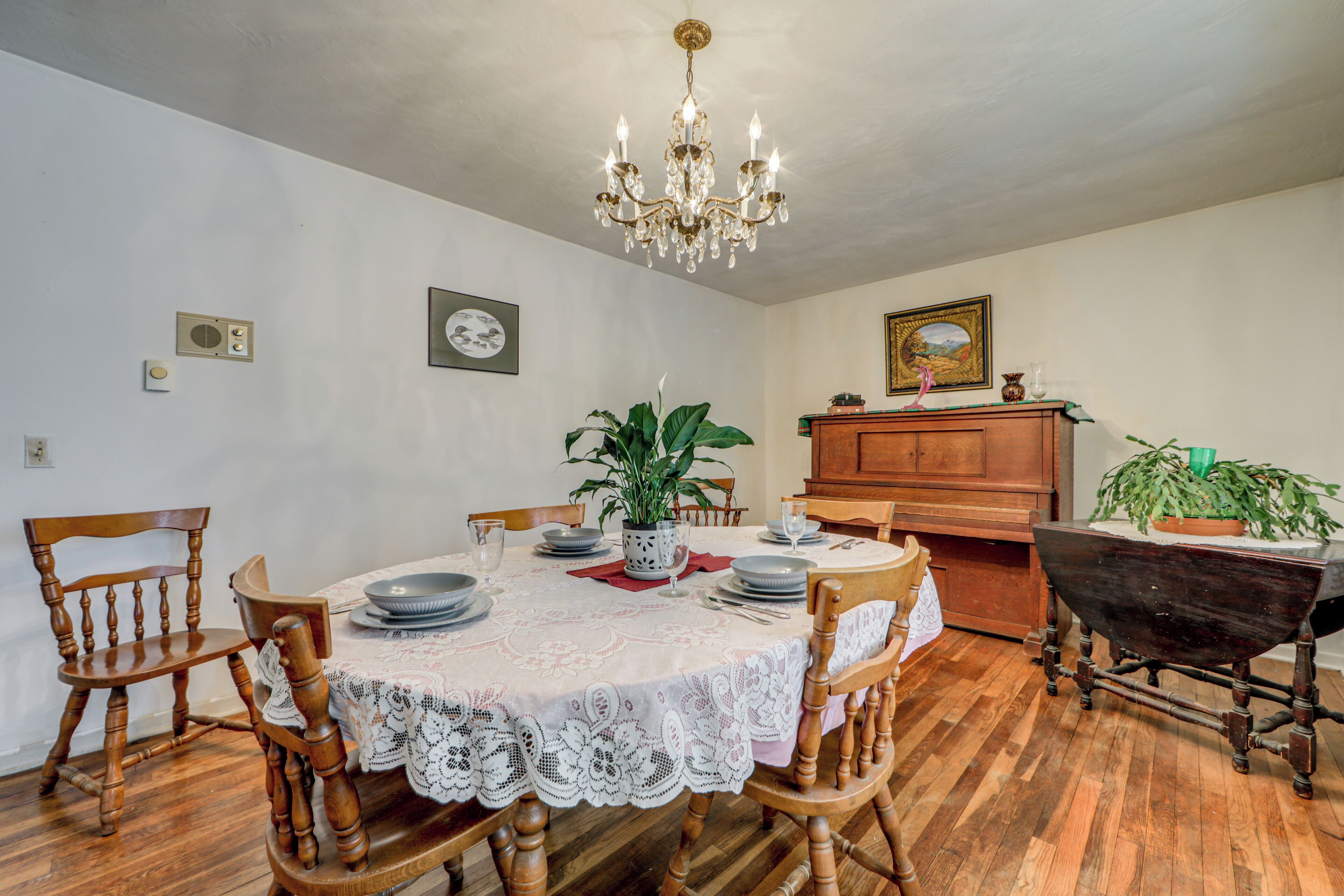 A dining room with a round table, chairs, a chandelier, a piano, and a potted plant.