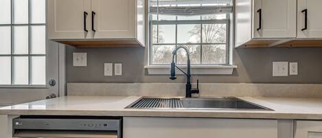 A kitchen with white cabinets, a stainless steel dishwasher, and a window with blinds.