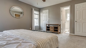 Une chambre à coucher comprenant un lit, un ventilateur de plafond, un miroir rond, une commode et une porte donnant sur une autre pièce.