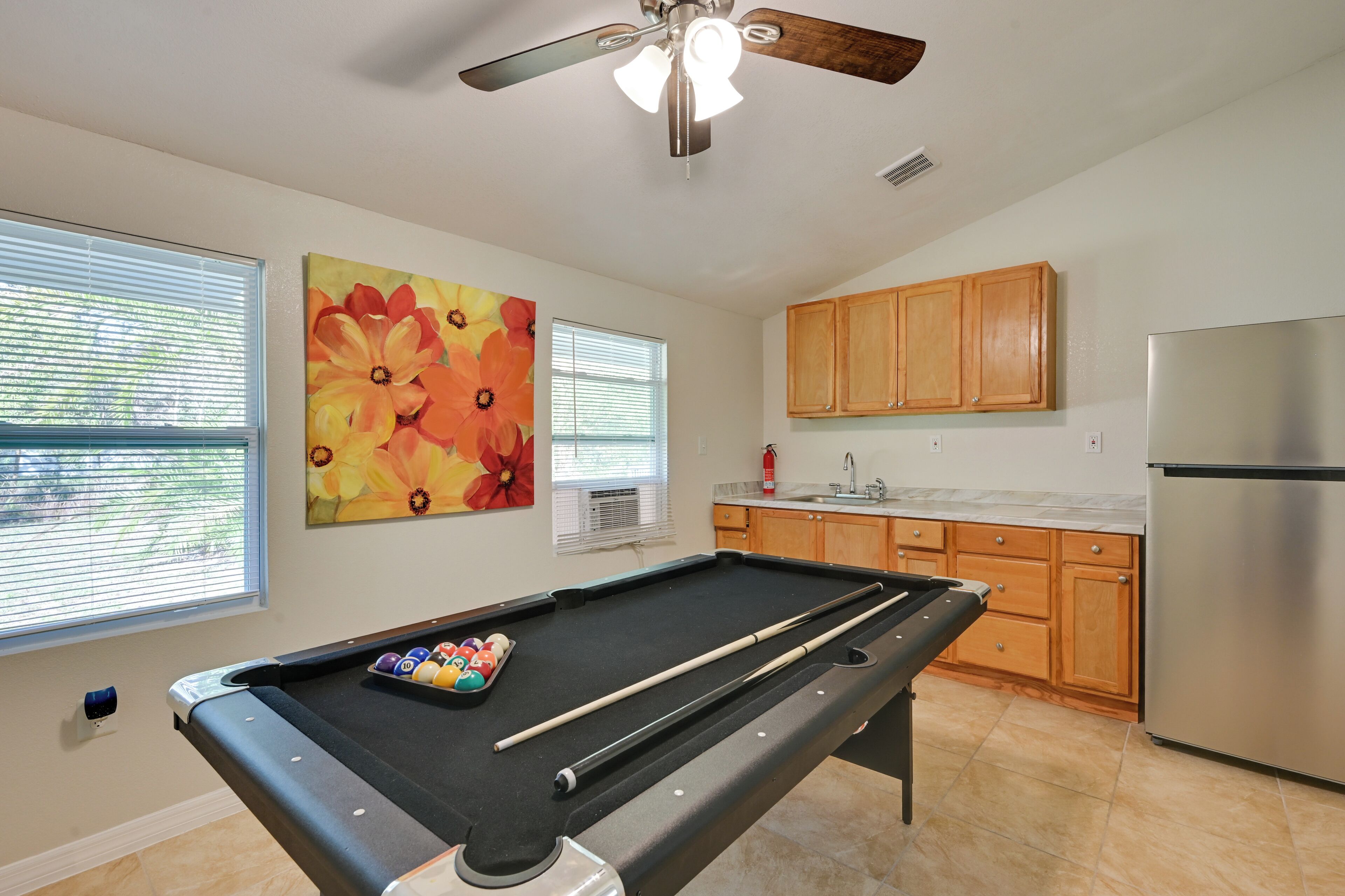 A pool table with balls, wooden cabinets, a stainless steel refrigerator, and a ceiling fan in a room with windows and a floral painting.