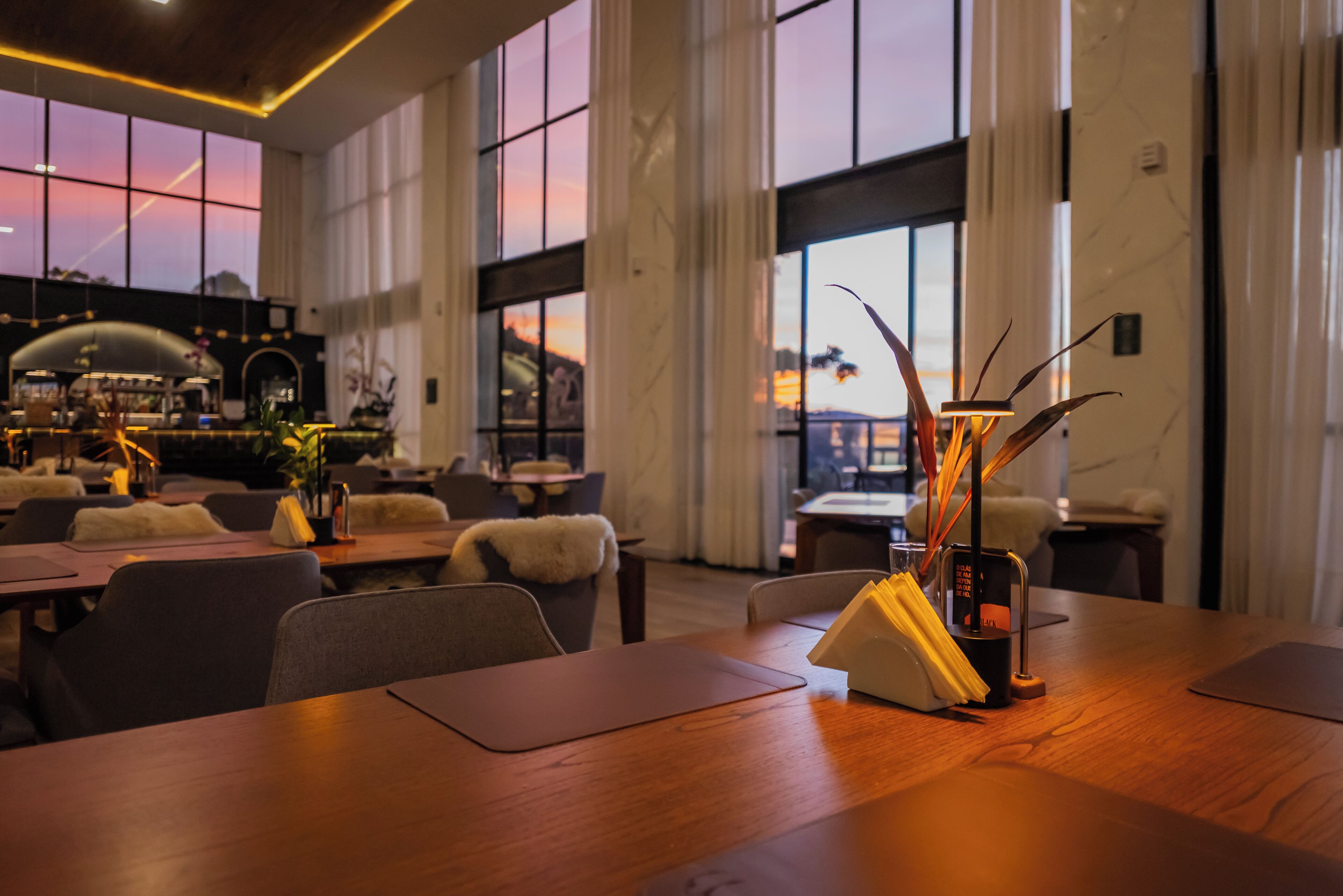Ein geräumiges Restaurant mit hohen Decken und großen Fenstern, das einen Blick auf den Sonnenuntergang bietet. Die Tische sind ordentlich mit Stühlen bestückt, und auf den Tischen stehen dekorative Pflanzen und Kerzenhalter.