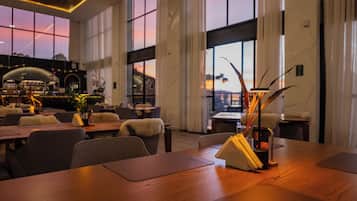 A spacious restaurant with high ceilings and large windows, offering a view of the sunset. Tables are neatly arranged with chairs, and there are decorative plants and a candle holder on the tables.