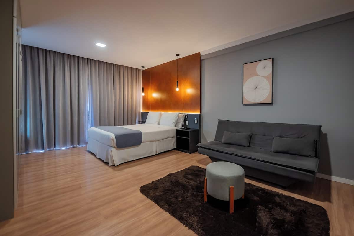 Ein modernes Hotelzimmer mit einem Bett, einer Couch, einem kleinen Hocker, einer Wandleuchte und einem gerahmten Bild.