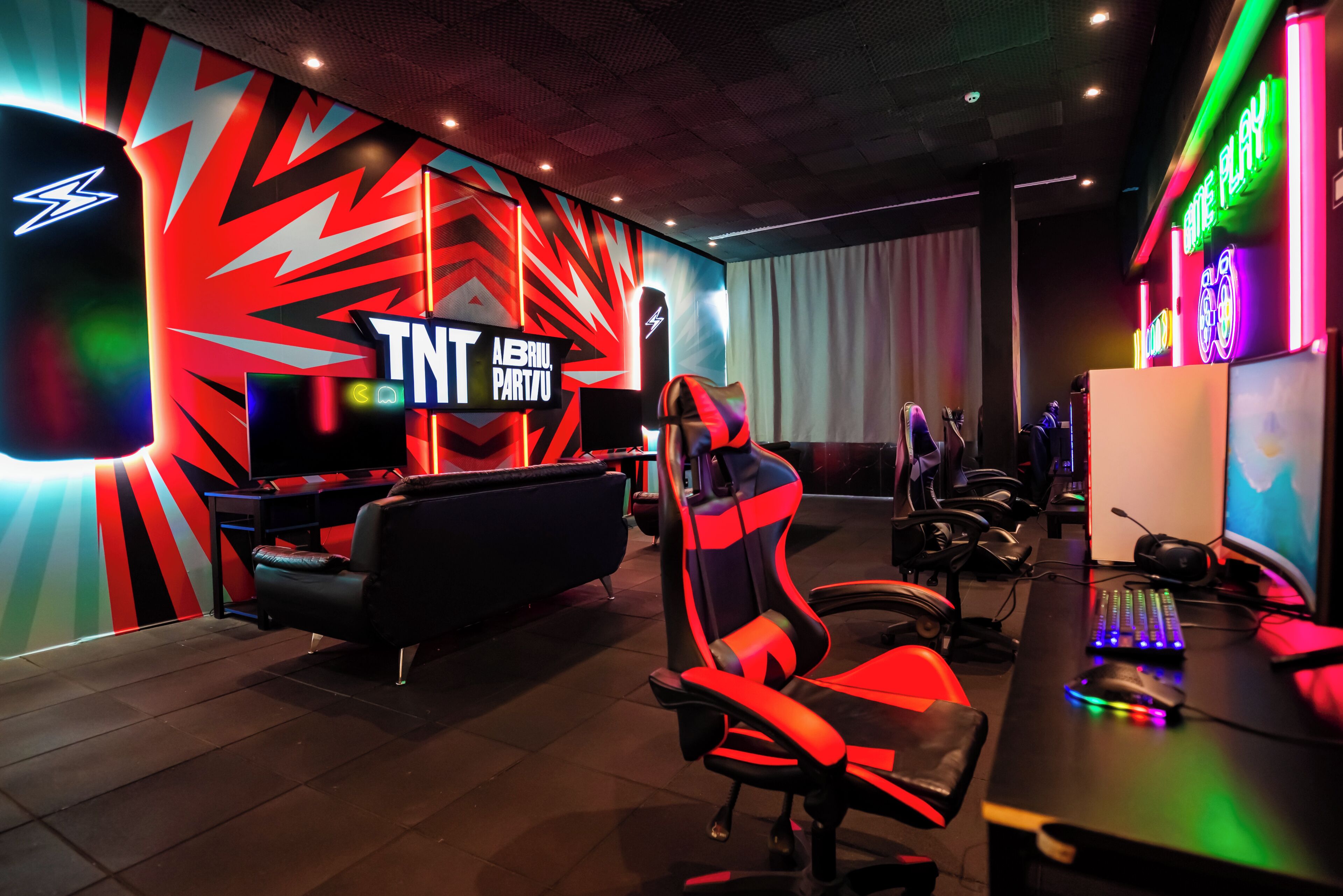 Een speelkamer met een neonbord, gamingstoelen en een bureau met een computer.