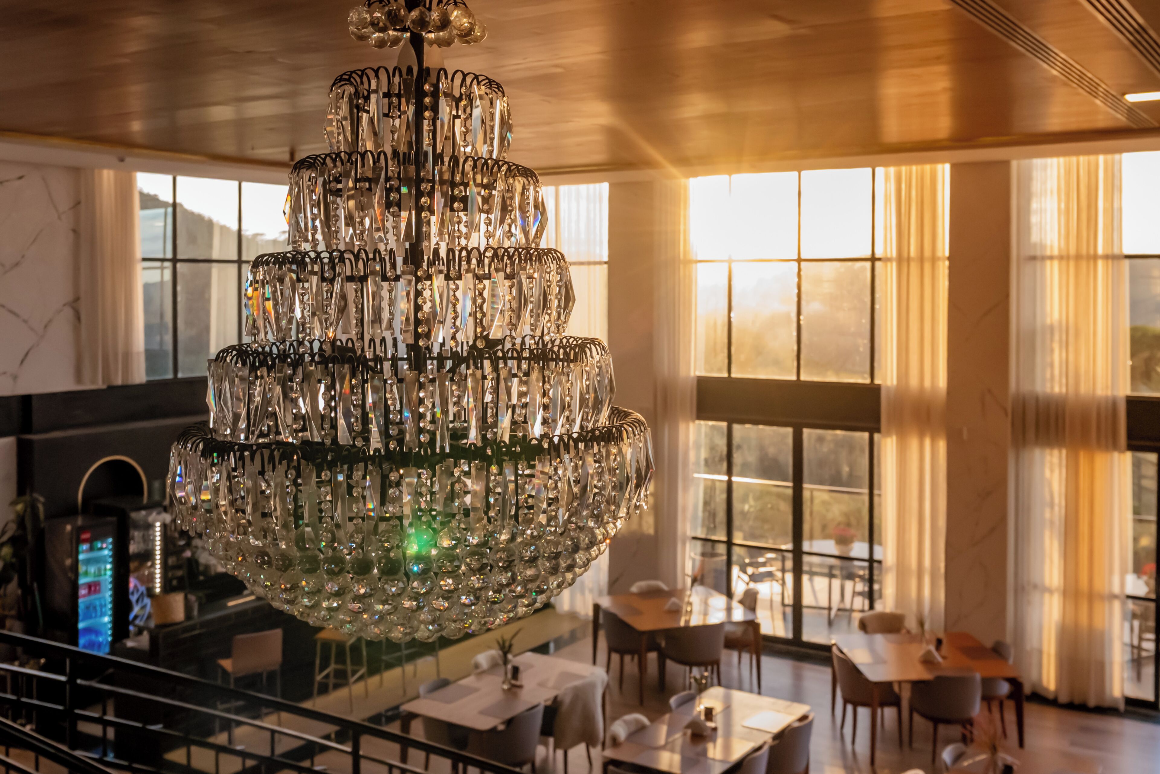 A chandelier in a dining area with tables and chairs, large windows, and a staircase.