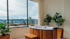 Une salle de bain moderne avec une baignoire en bois, un coin salon et une grande fenêtre donnant sur les montagnes.