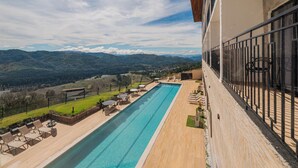 Une longue piscine, des chaises longues et des tables, une terrasse en bois et une vue sur les montagnes.
