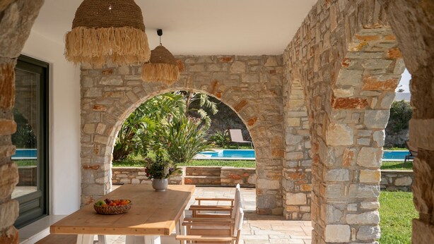 An outdoor patio with a stone archway, a wooden table with chairs, and a view of a swimming pool.