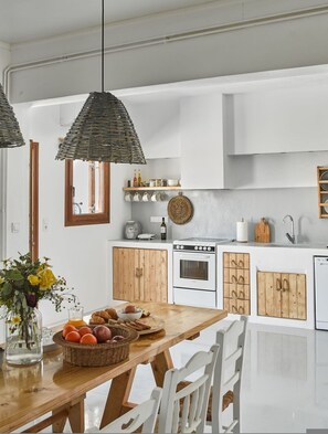 A modern kitchen with white cabinets, a wooden dining table, hanging wicker pendant lights, and a view of the outdoors. - Maria Seaview Villa near Naousa (Paros)