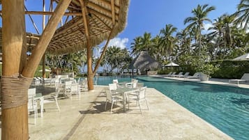 Un área de piscina con sillas de descanso, una estructura con techo de paja y palmeras.