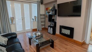 Una sala de estar con sofá de cuero negro, una mesa de centro de vidrio, una chimenea, un televisor montado en la pared y un ventanal con cortinas.