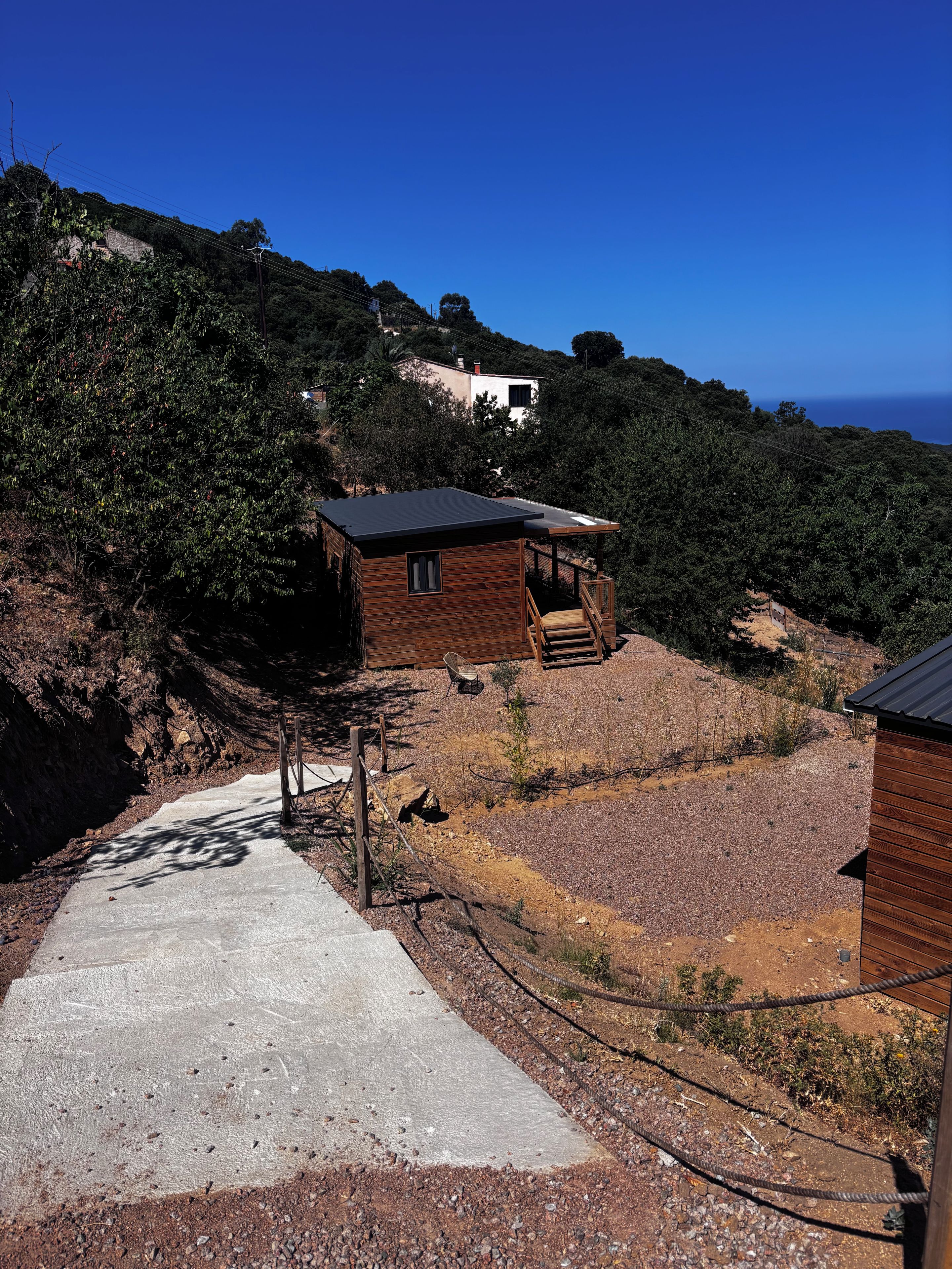 Une cabane en bois avec un toit en pente, un chemin de béton menant à celle-ci, et une vue sur une colline couverte d’une végétation dense.
