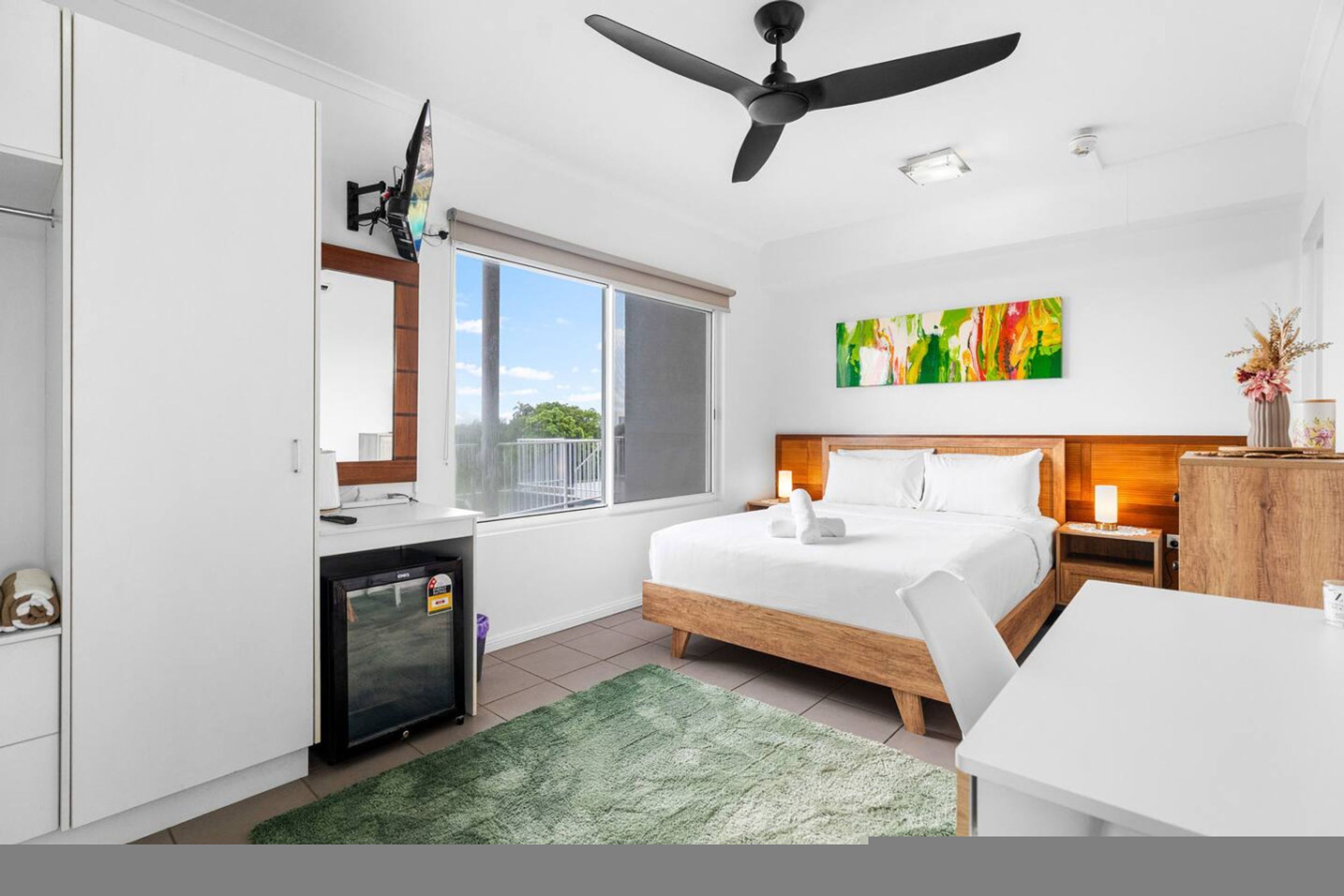 A hotel room with a bed, a desk, a refrigerator, a ceiling fan, and a large window.