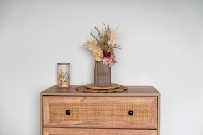 Een nachtkastje van hout met een gevlochten lade, een vaas met gedroogde bloemen en een cilindervormig doosje met bloemendesign.
