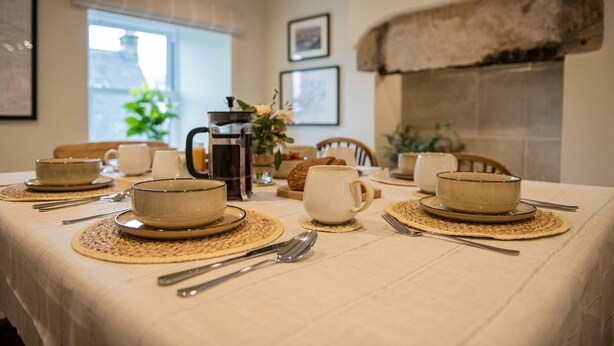 A dining table set with bowls, cups, and cutlery, with a rustic fireplace and framed pictures in the background. - Charming Countryside Cottage Yorkshire Dales (Richmond)