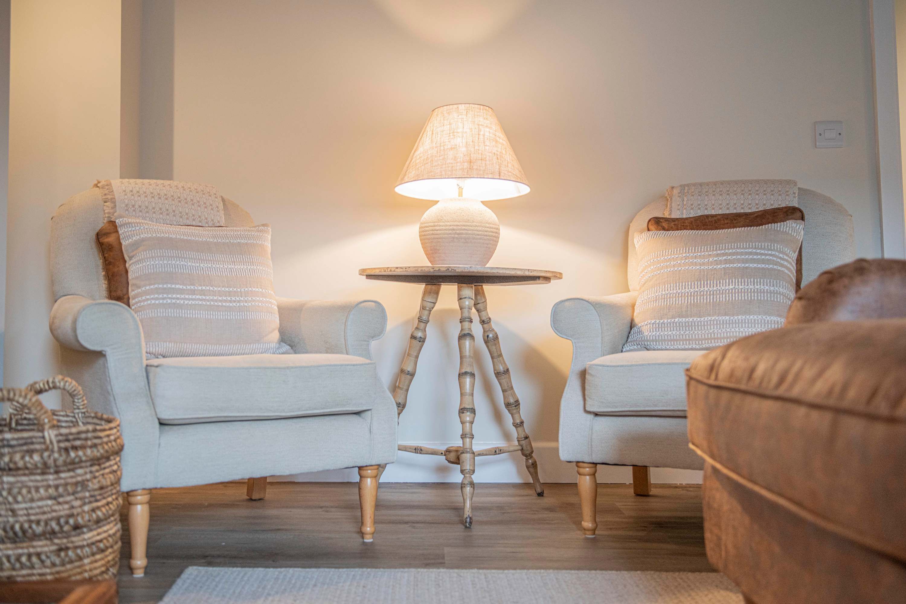 A room with two armchairs, a round table with a lamp, a woven basket, and a brown sofa.
