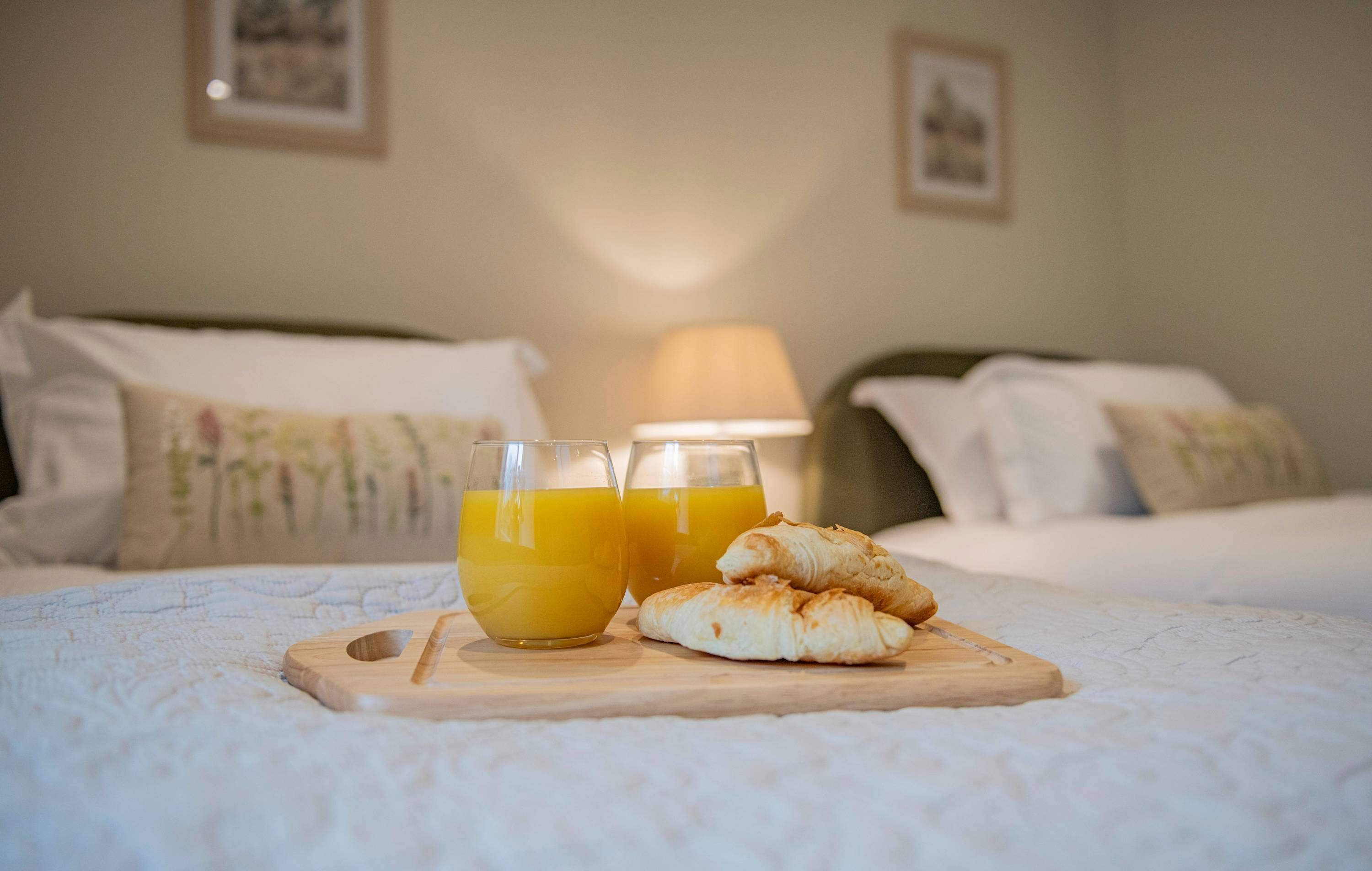 Una cama con almohadas, una mesita de noche con dos vasos de jugo de naranja y una tabla de madera con croissants.