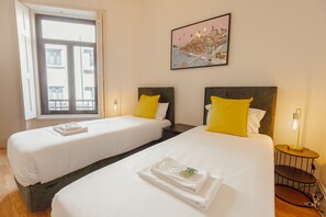 Habitación de hotel con dos camas, una ventana con vistas a edificios, un cuadro en la pared y una mesita de noche con una lámpara.
