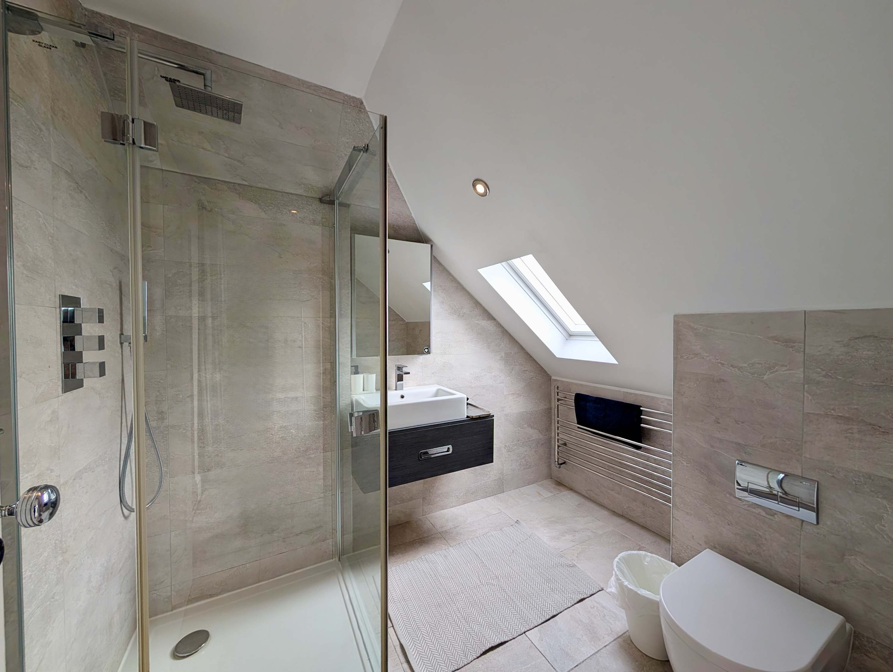 A modern bathroom with a glass-enclosed shower, a skylight, a white sink, and a toilet.