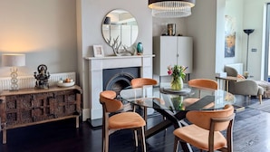Ein modernes Wohnzimmer mit Kamin, einem runden Spiegel, einer Chandelier-Leuchte und einem Esstisch mit Glasplatte und orangefarbenen Stühlen.