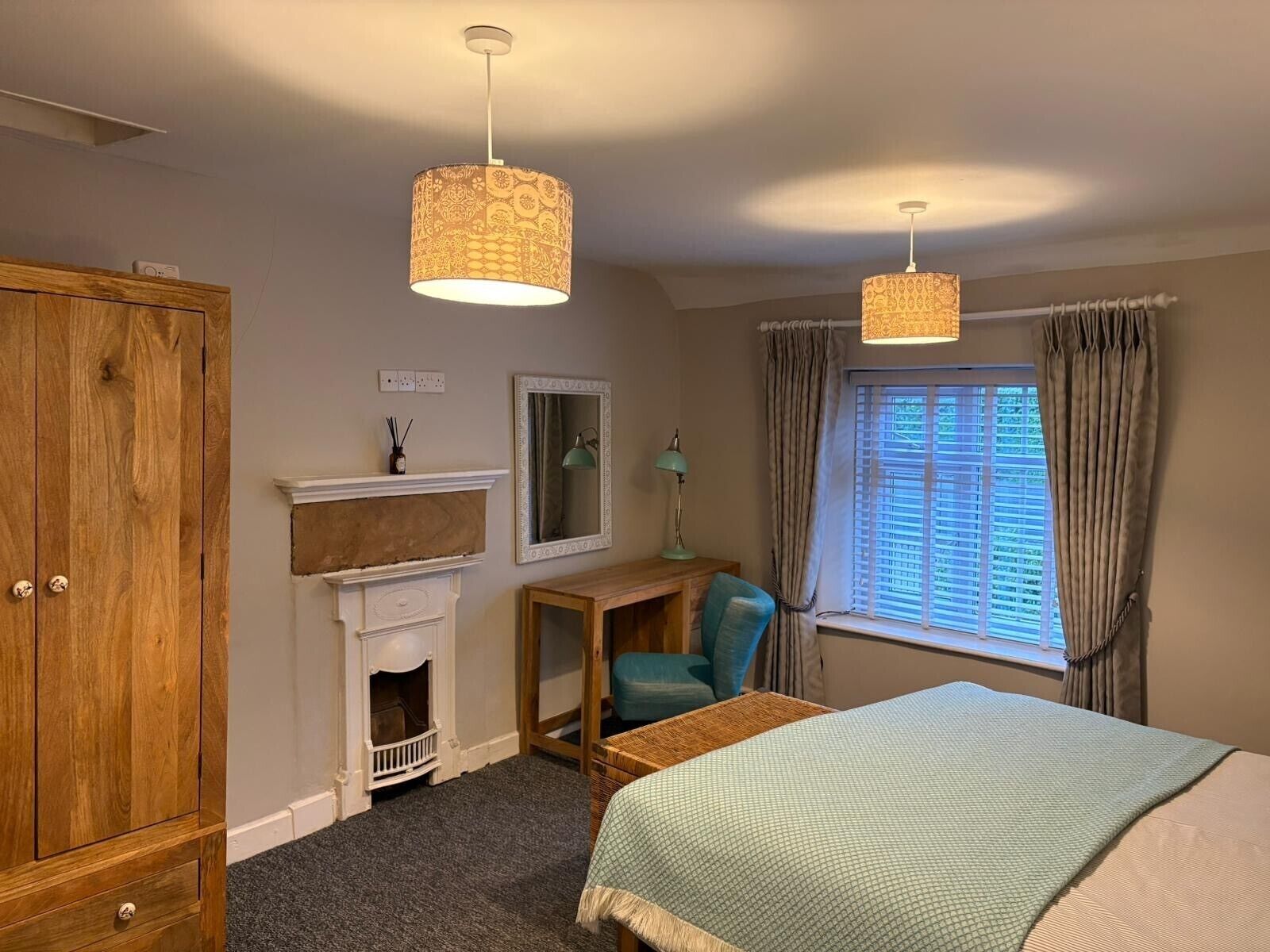 A bedroom with a wooden wardrobe, a fireplace, a desk with a lamp, a bed, and a window with curtains.