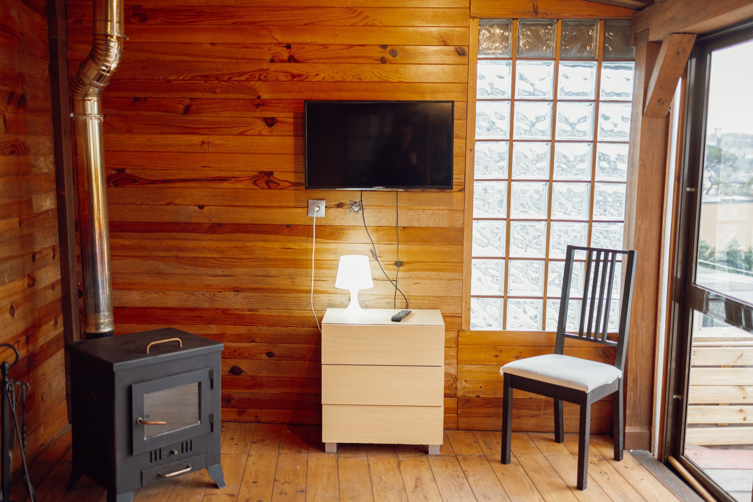 A wooden cabin interior with a wood stove, a TV mounted on the wall, a bedside table with a lamp, and a window with a view.