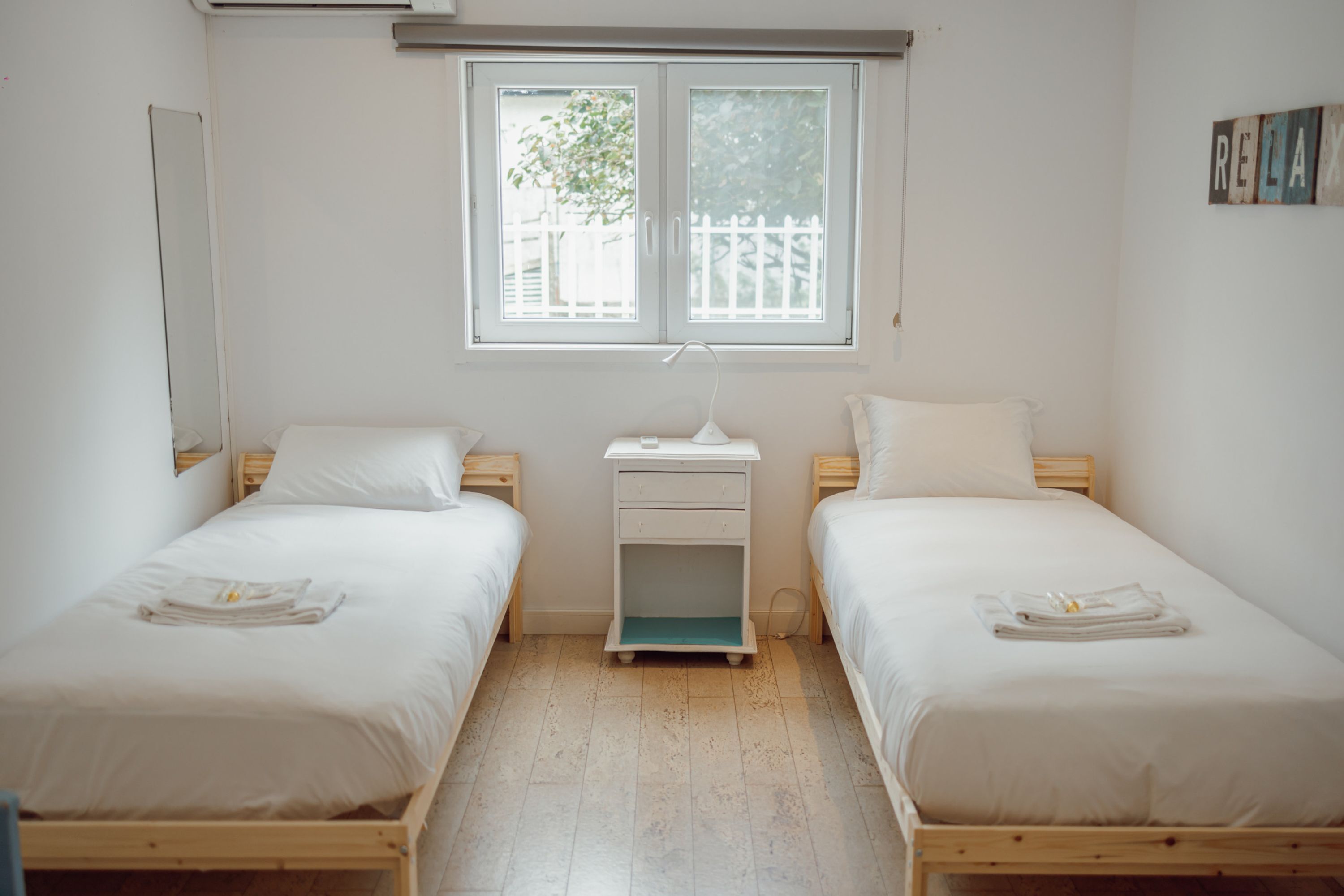 A twin room with two single beds, a small bedside table, a nightstand, and a window with a view of a white fence.