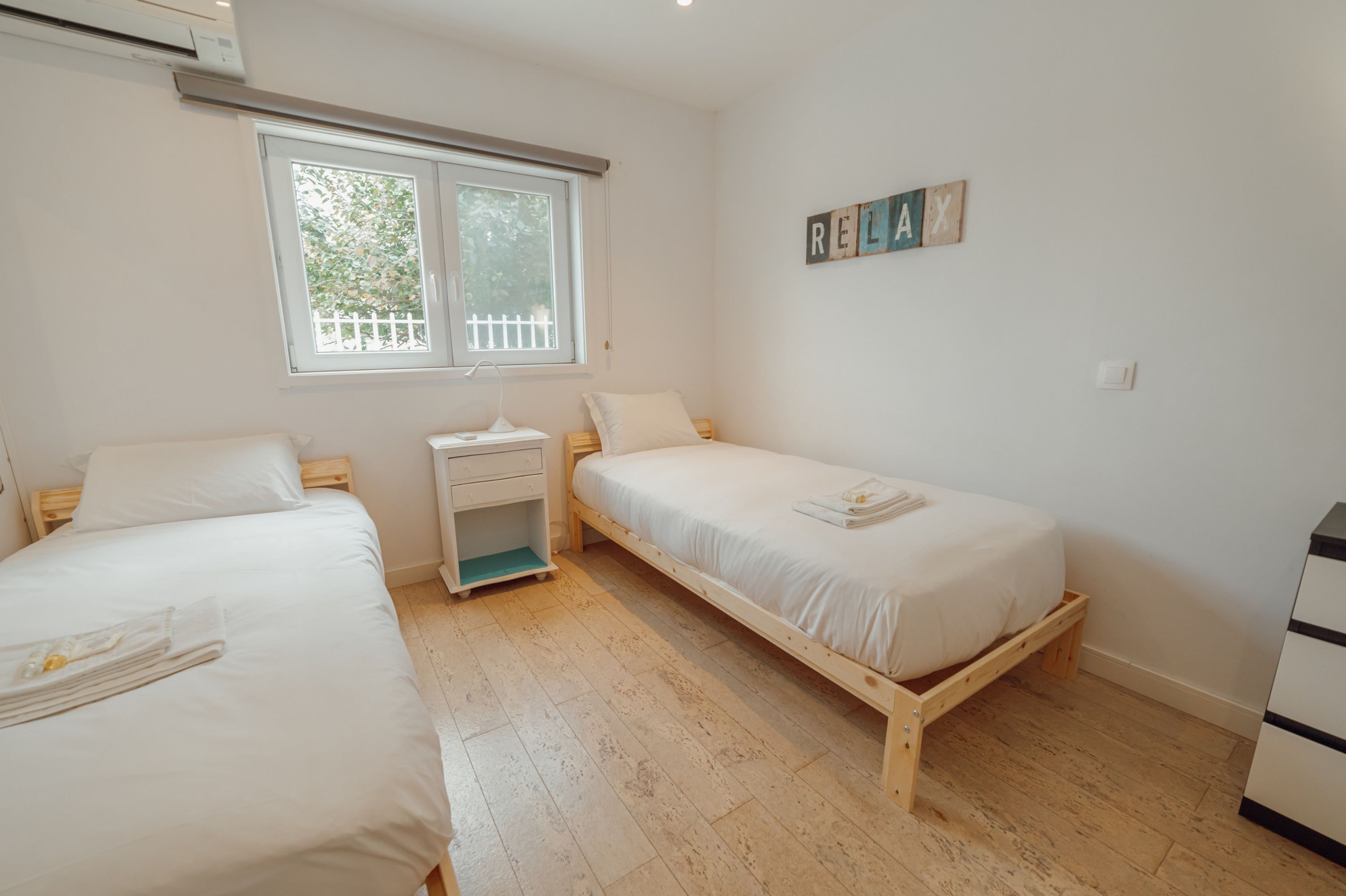 A bedroom with two single beds, a bedside table, a nightstand, a window with a view of greenery, and a wall-mounted word puzzle.
