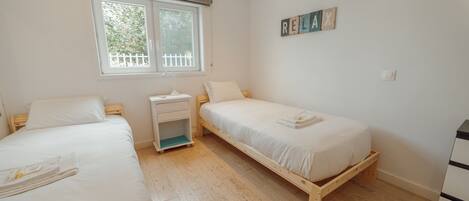 A bedroom with two single beds, a bedside table, a nightstand, a window with a view of greenery, and a wall-mounted word puzzle.