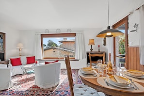 Une pièce avec une table à manger dressée pour un repas, un canapé avec des coussins rouges, un tapis à motifs et une vue sur un bâtiment à l’extérieur.