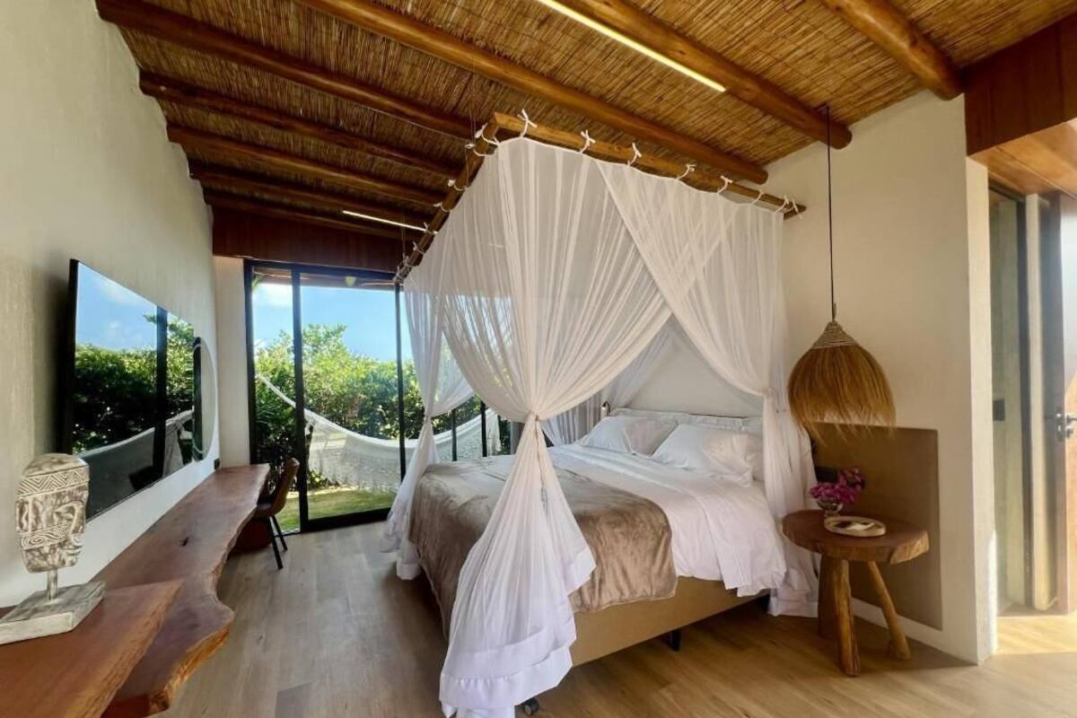 Un dormitorio con cama con dosel, un banco de madera, un televisor y vistas al exterior con vegetación.