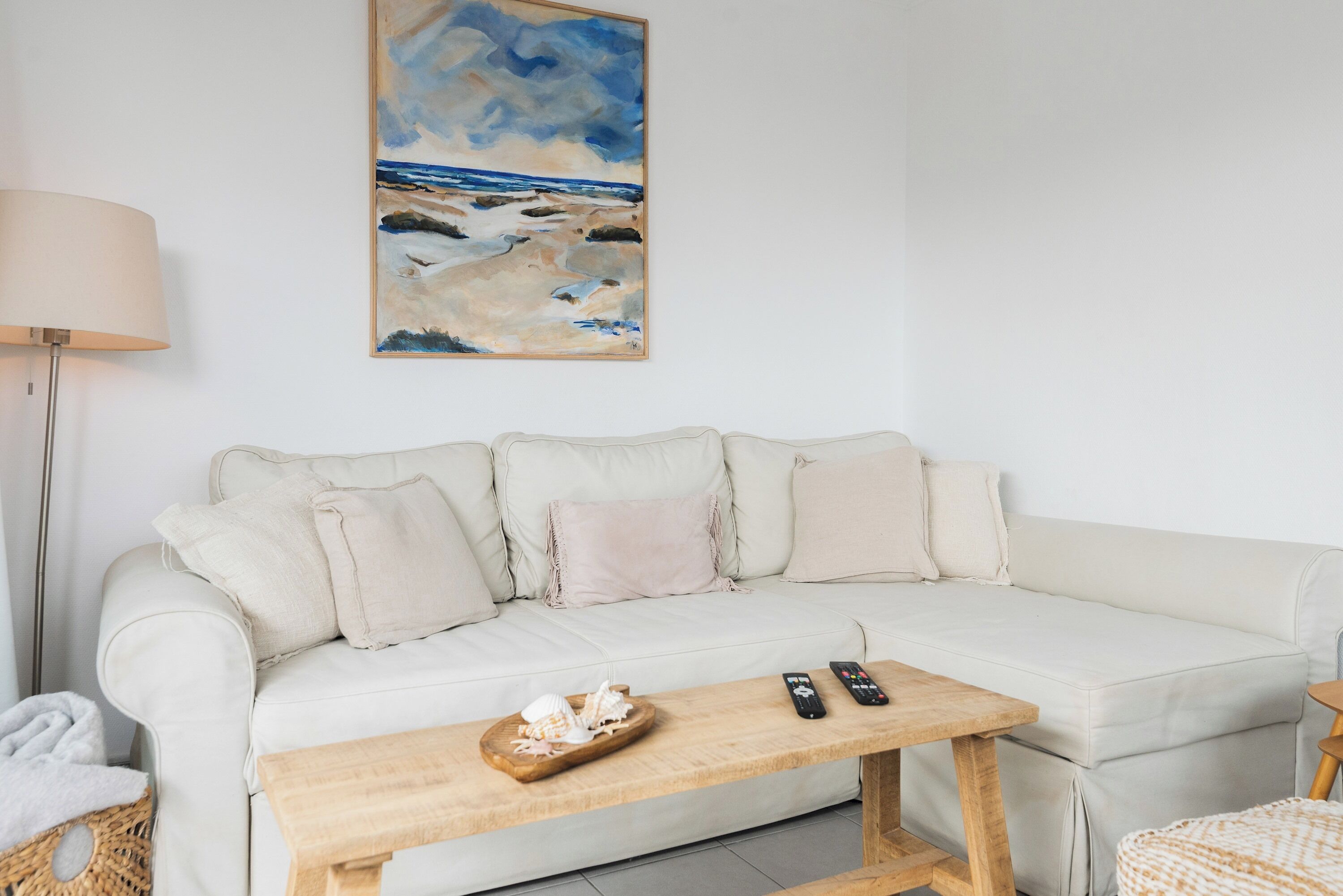 Ein weißes Sofa mit Kissen, ein Couchtisch aus Holz mit einem dekorativen Gegenstand, einer Fernbedienung und ein Gemälde mit Strandmotiv an der Wand.