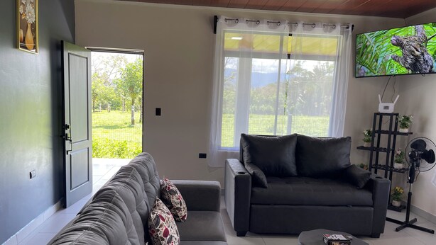 A living room with a grey sofa, a dark armchair, a coffee table, a TV, and a window with a view of greenery. - Charming 2-bedroom villa with AC in peaceful  de La Fortuna, San Carlos (San Carlos)