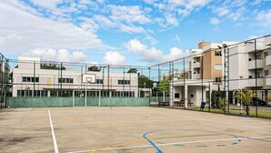An outdoor basketball court with a chain-link fence, a basketball hoop, and a building with multiple windows and balconies in the background. - Fully Equipped Apartments in the Condo With Pool in Canasvieiras - RPC (Florianópolis)