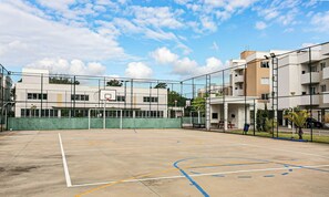 An outdoor basketball court with a chain-link fence, a basketball hoop, and a building with multiple windows and balconies in the background. - Fully Equipped Apartments in the Condo With Pool in Canasvieiras - RPC (Florianópolis)