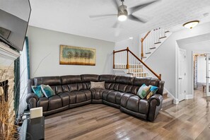 A living room with a sectional sofa, a painting on the wall, a staircase, and a ceiling fan. - Fabulous 4BR Home, Office, Game Room & Deck/Huge Lawn (Conyers)