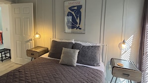 Une chambre avec un grand lit, une table de chevet, une commode, une œuvre d’art encadrée au mur et un luminaire suspendu.