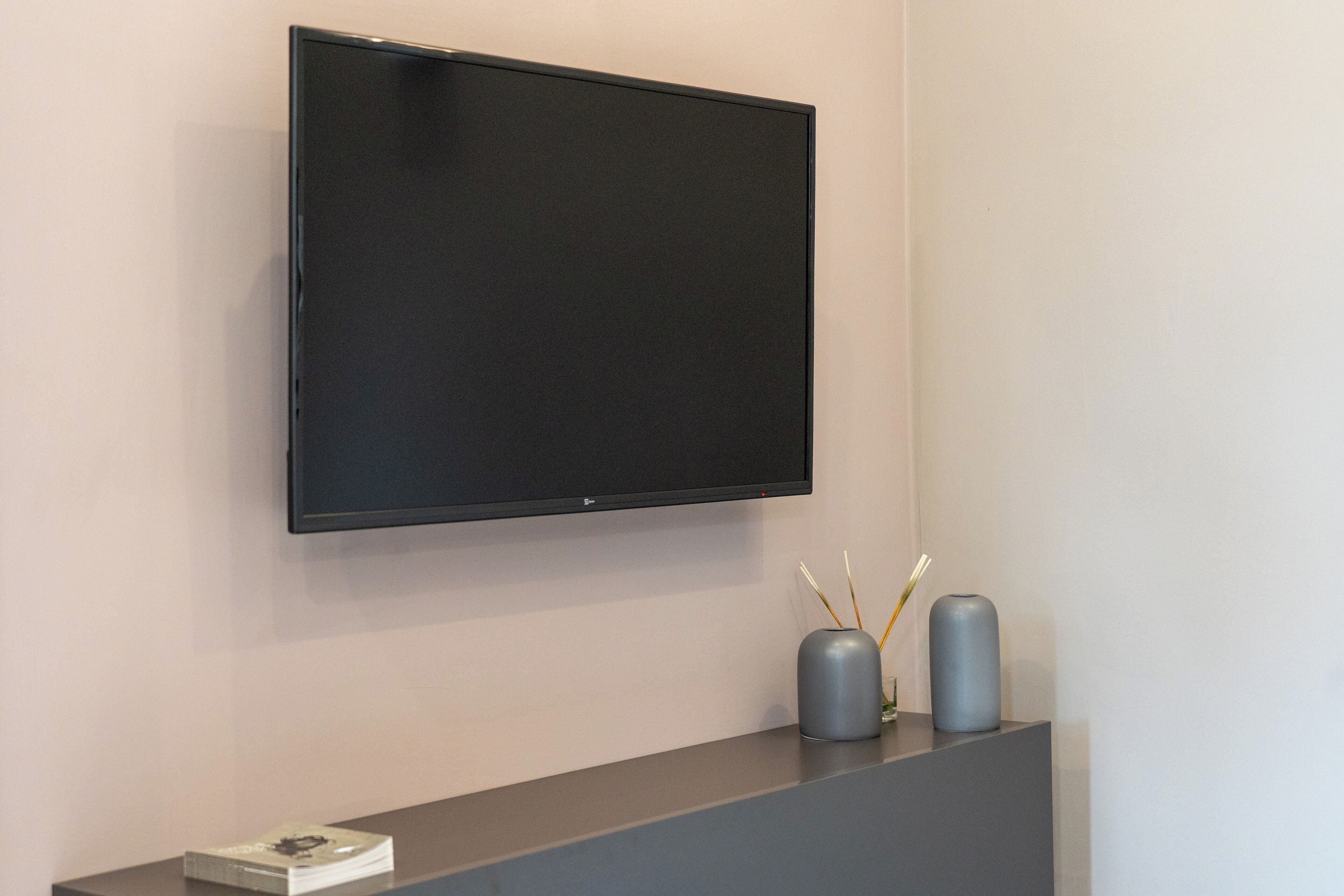 Un televisor de pantalla plana montado en la pared, una consola oscura con dos jarrones grises y un objeto blanco pequeño sobre la mesa.