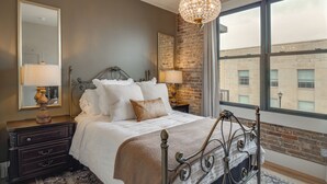 A bedroom with a large bed, a dresser, a mirror, a chandelier, and a view of a building through a window. - Historic Lawrenceburg Loft: Balcony + Square Views (Lawrenceburg)
