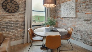 A room with a brick wall, a round table with chairs, a clock, a chandelier, and a window with a view. - Historic Lawrenceburg Loft: Balcony + Square Views (Lawrenceburg)