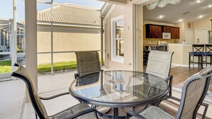 Een tafel met glazen blad en stoelen in een overdekt buitengebied, grenzend aan een huis waar een keuken door een open deur zichtbaar is.