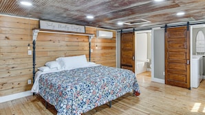 Quarto com cabeceira de madeira, cama com colcha floral, ar-condicionado fixado na parede e porta deslizante para um banheiro.