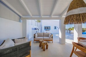 Ein überdachter Außenbereich mit einer Couch, Stühlen und einem Tisch, mit Blick auf einen Pool und ein Gebäude mit blauen Akzenten.